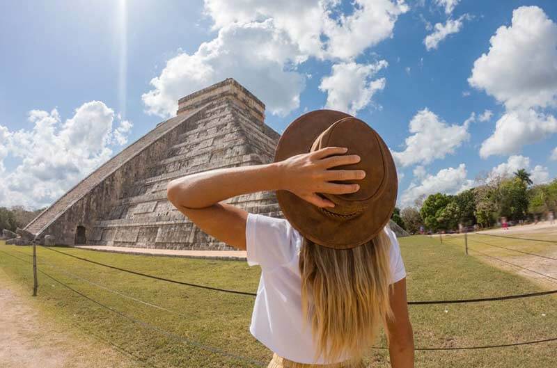 Woman with hat facing Chichen Itza pyramid in Mexico on a sunny day